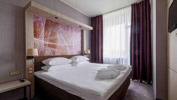 Habitación de hotel con una cama grande, dos lámparas de noche, televisión y vistas al exterior a través de cortinas translúcidas.