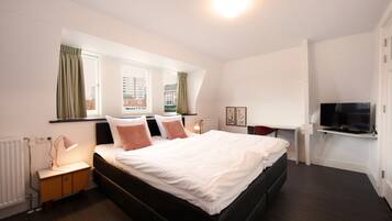 A modern hotel room with a large bed, a bedside table with a lamp, a flat-screen TV mounted on the wall, and a window with curtains.
