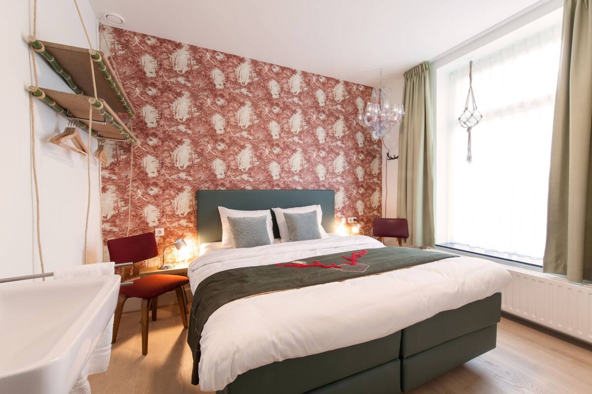 Une chambre à coucher comprenant un lit, deux tables de chevet, une chaise, un bureau, un luminaire suspendu et une fenêtre avec des rideaux.