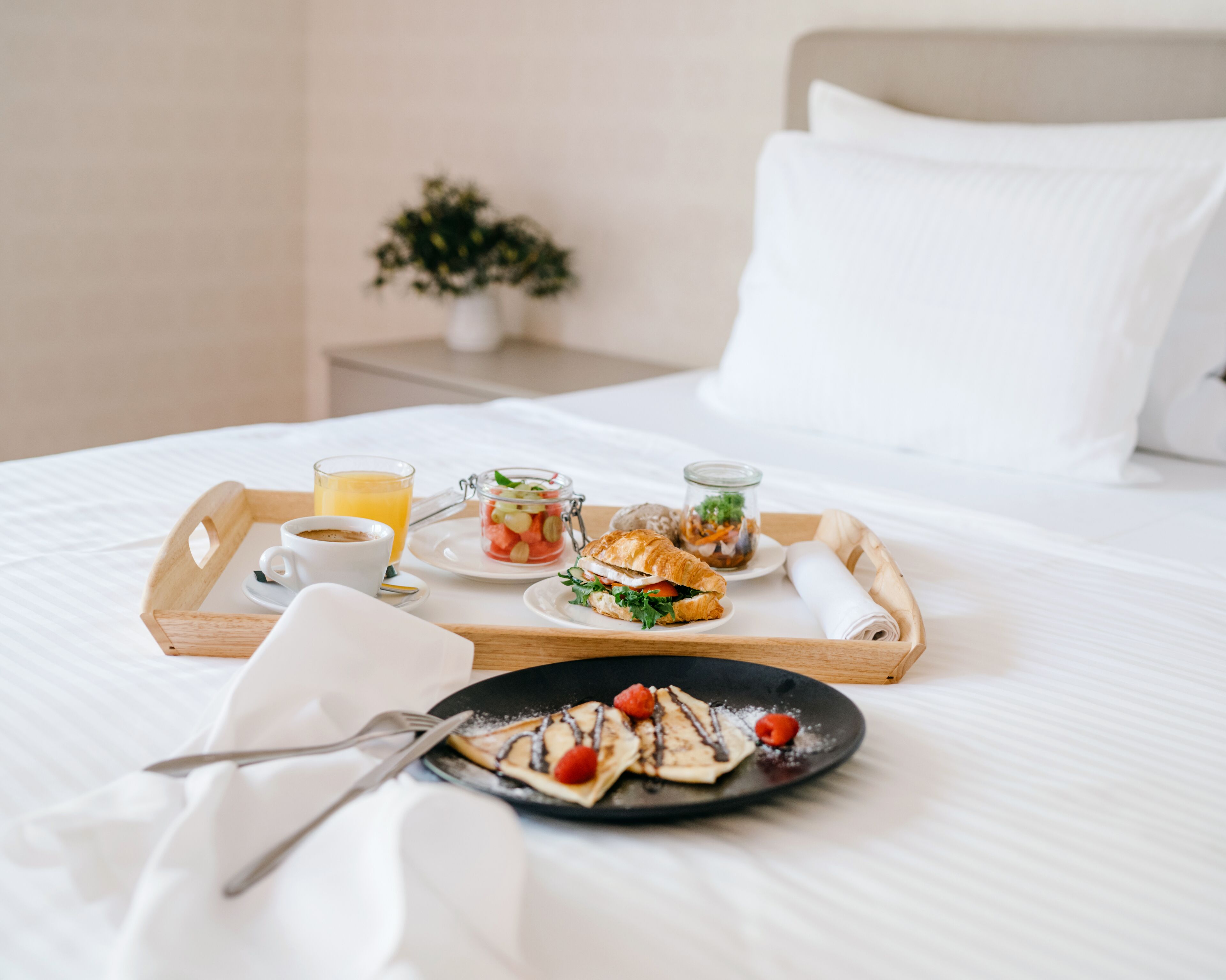Una bandeja de desayuno ordenada sobre una cama con una almohada blanca y una pequeña planta en maceta al fondo.