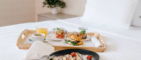 Una bandeja de desayuno ordenada sobre una cama con una almohada blanca y una pequeña planta en maceta al fondo.
