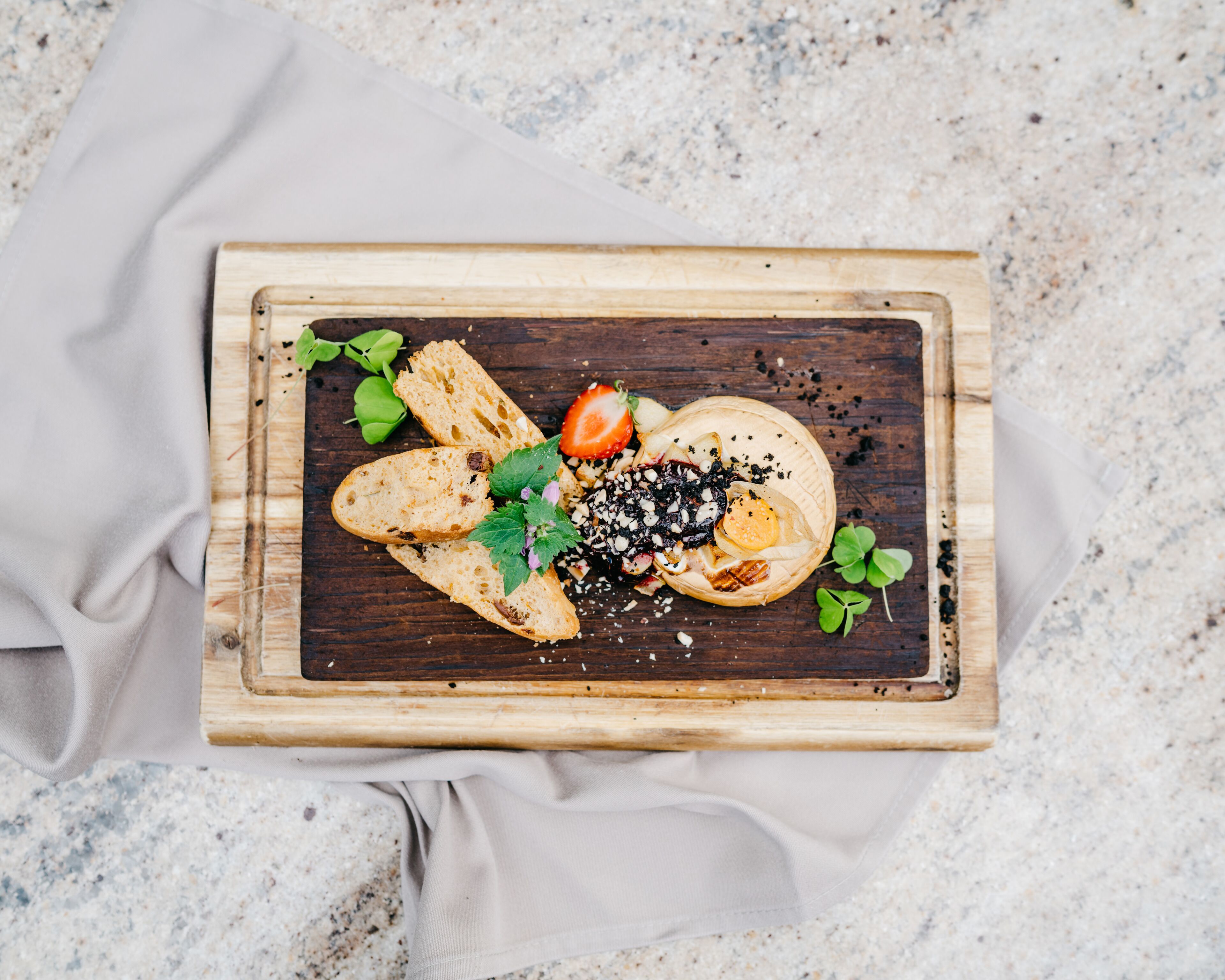 A wooden board with food items including bread, a strawberry, and a cheese wheel, placed on a speckled surface.