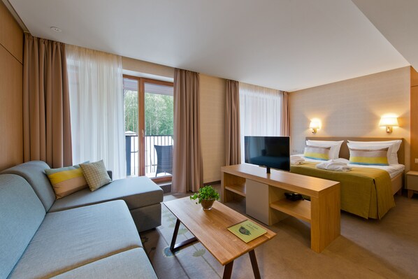 Una habitación de hotel moderna con sofá, cama, televisión, una mesita con una planta y un balcón con cortinas.