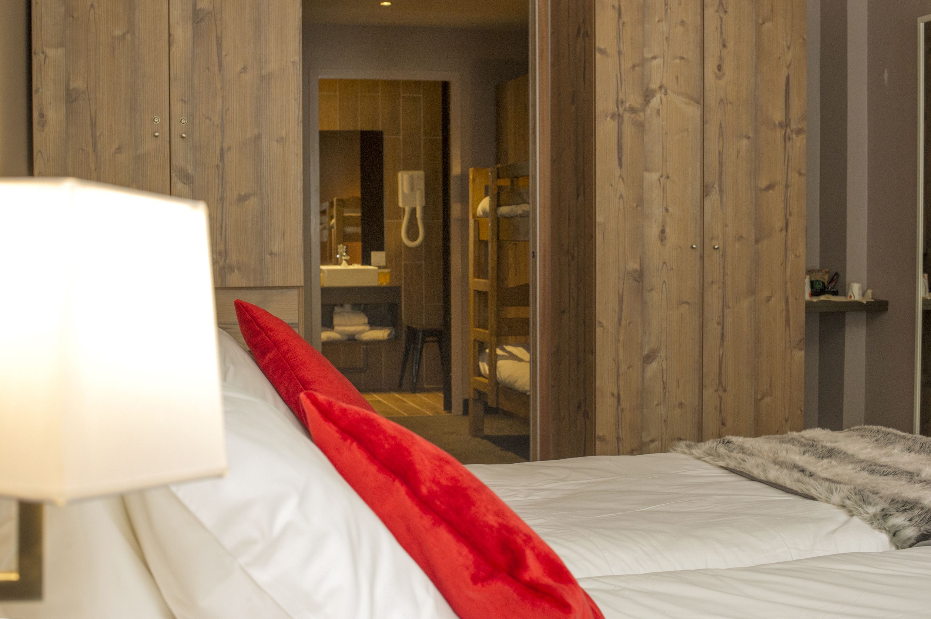 Ett hotellrum med en säng, en röd kudde, en lampa och en garderob i trä.
