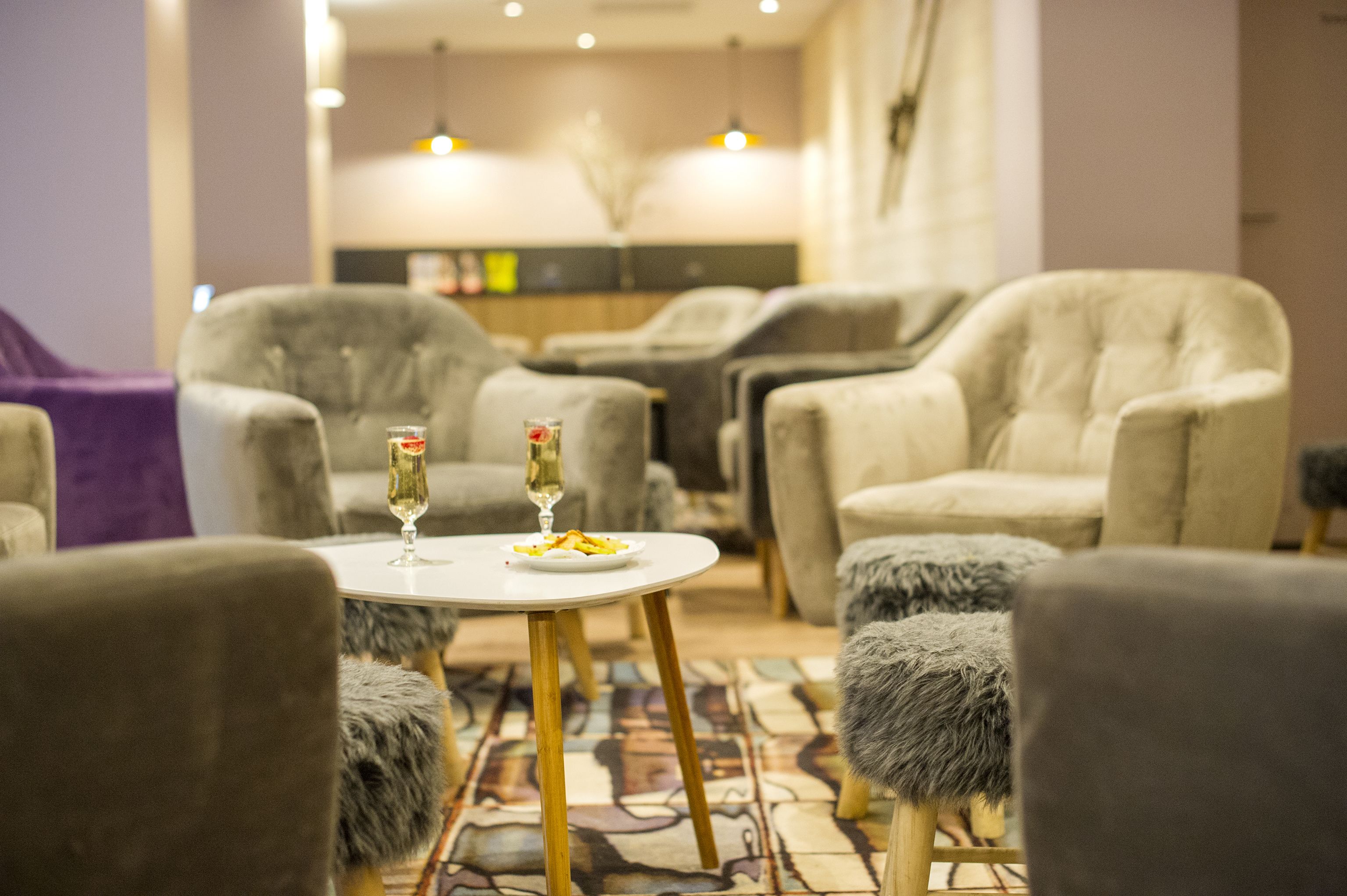 A modern lounge area with plush armchairs, a round table with a glass of wine and a plate of food, and a patterned rug.