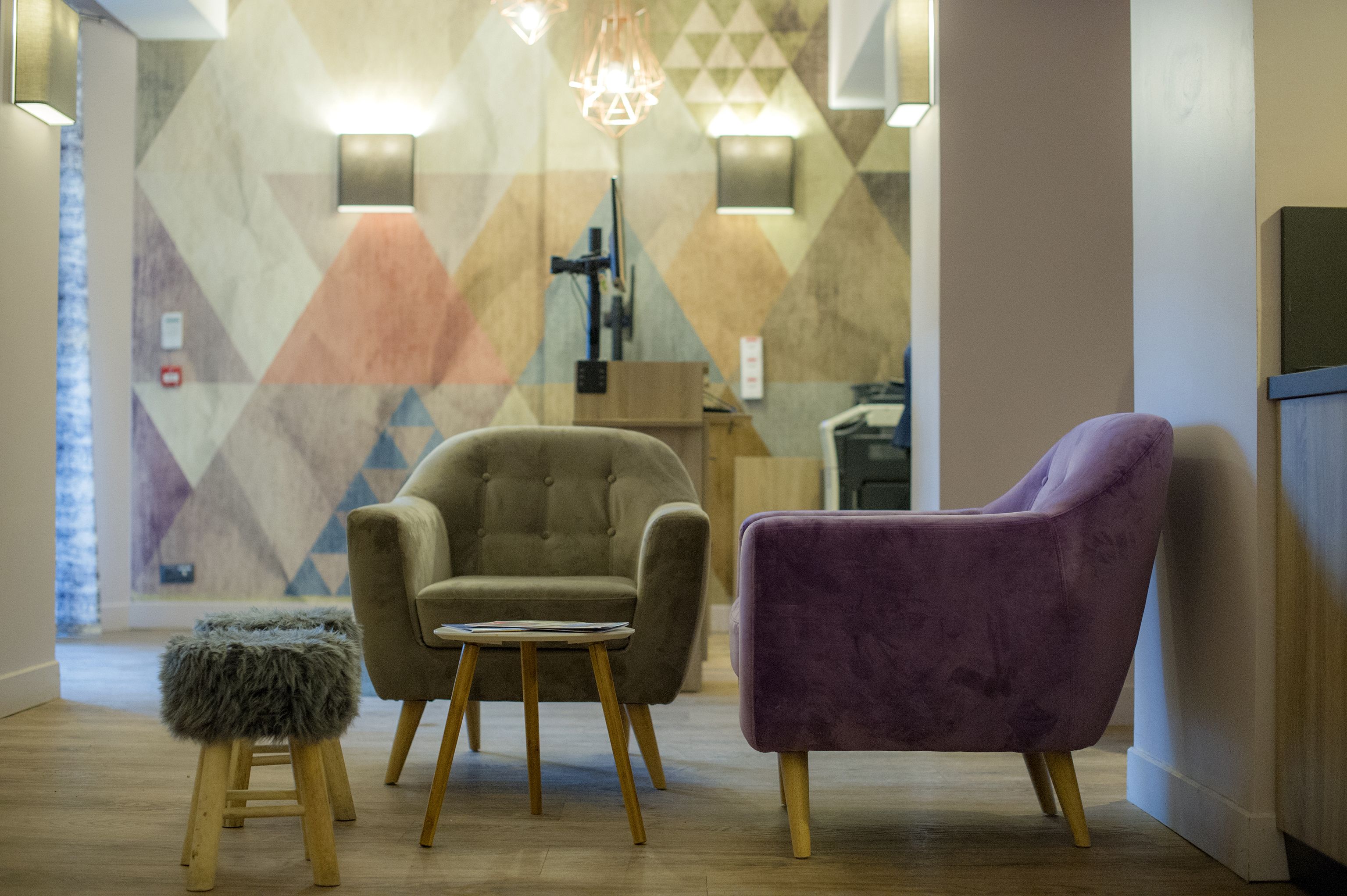 A modern lobby with a geometric patterned wall, a beige armchair, a purple armchair, and a small wooden stool.