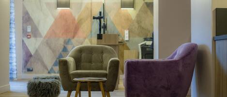 A modern lobby with a geometric patterned wall, a beige armchair, a purple armchair, and a small wooden stool.