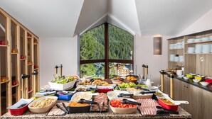 A buffet setup with various food items and utensils, a large window with a forest view, and wooden shelves with glassware. - SOWELL Family Valfréjus (Modane)