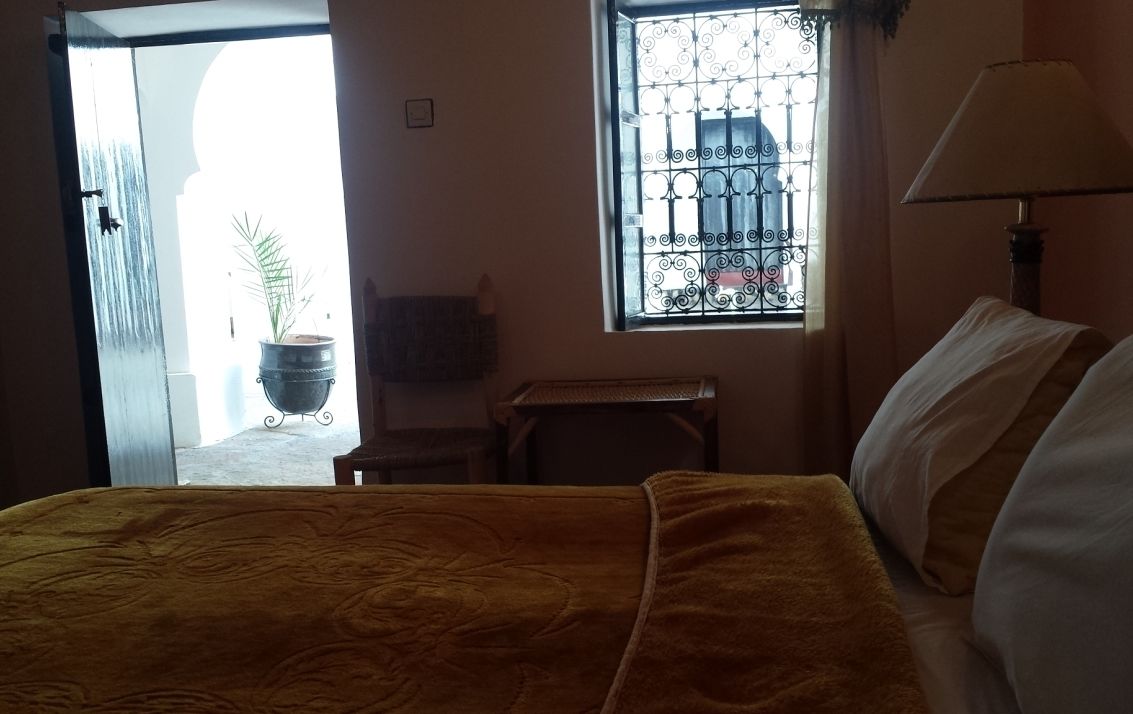 A room with a bed, a window with metal bars, a lamp, and a door leading outside.