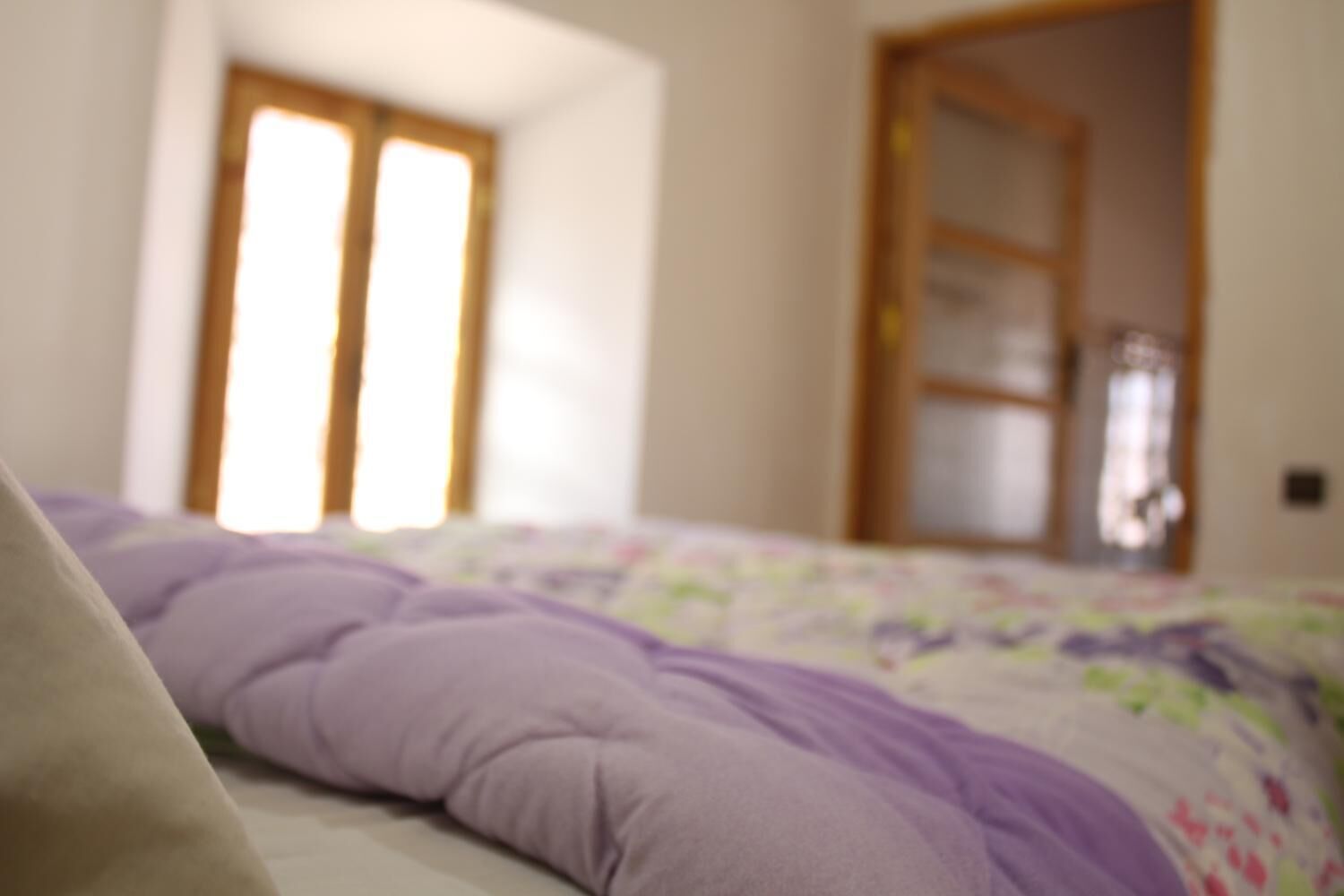 A bed with a purple blanket and a floral bedspread in a room with a wooden door and a window with blinds.