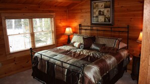 A wooden cabin bedroom with a bed, two bedside lamps, a window with blinds, and a framed picture on the wall. - Lake Parlin Lodge & Cabins (Jackman)