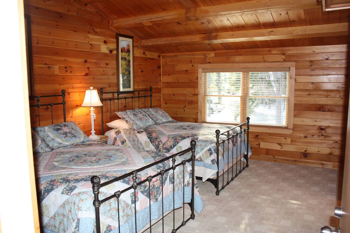 A wooden cabin bedroom with two beds, a window, and a nightstand with a lamp.