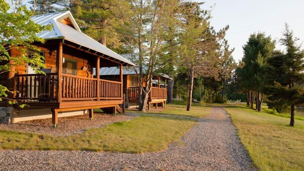 A cabin with a wooden deck and a metal roof, surrounded by trees and a gravel path. - Lake Parlin Lodge & Cabins (Jackman)