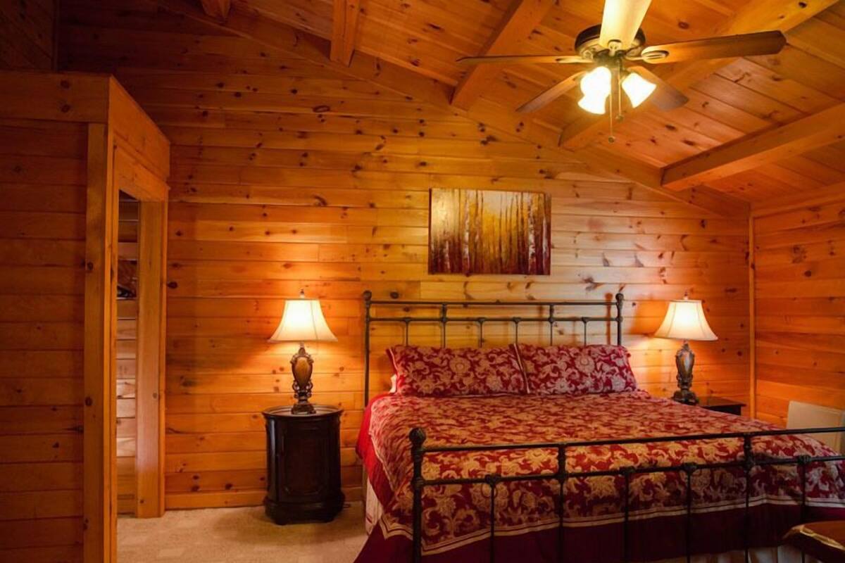 A wooden cabin bedroom with a bed, two bedside lamps, a ceiling fan, and a wooden door.