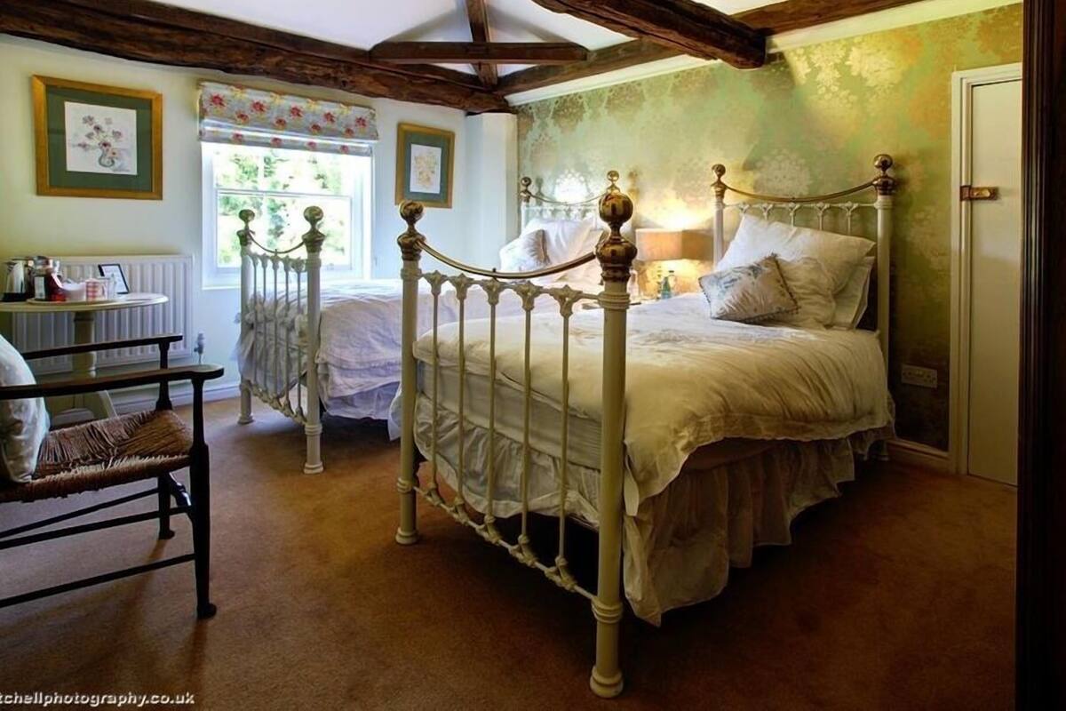 A bedroom with a four-poster bed, a wooden chair, a radiator, and a window with floral curtains.