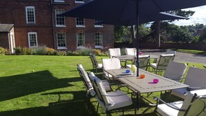 Eine Terrasse im Freien mit Tischen und Stühlen, ein Backsteinhaus im Hintergrund und eine Rasenfläche.