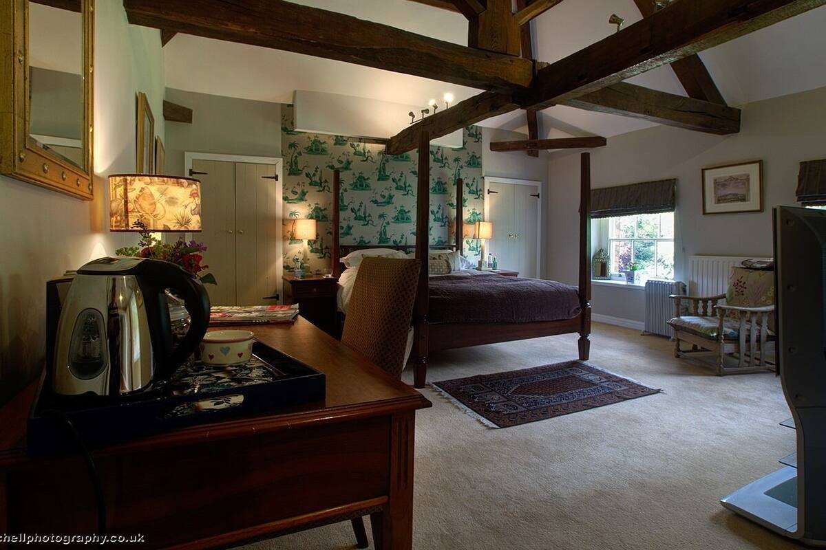 A bedroom with a four-poster bed, a wooden desk with a kettle, a chair, and a radiator.