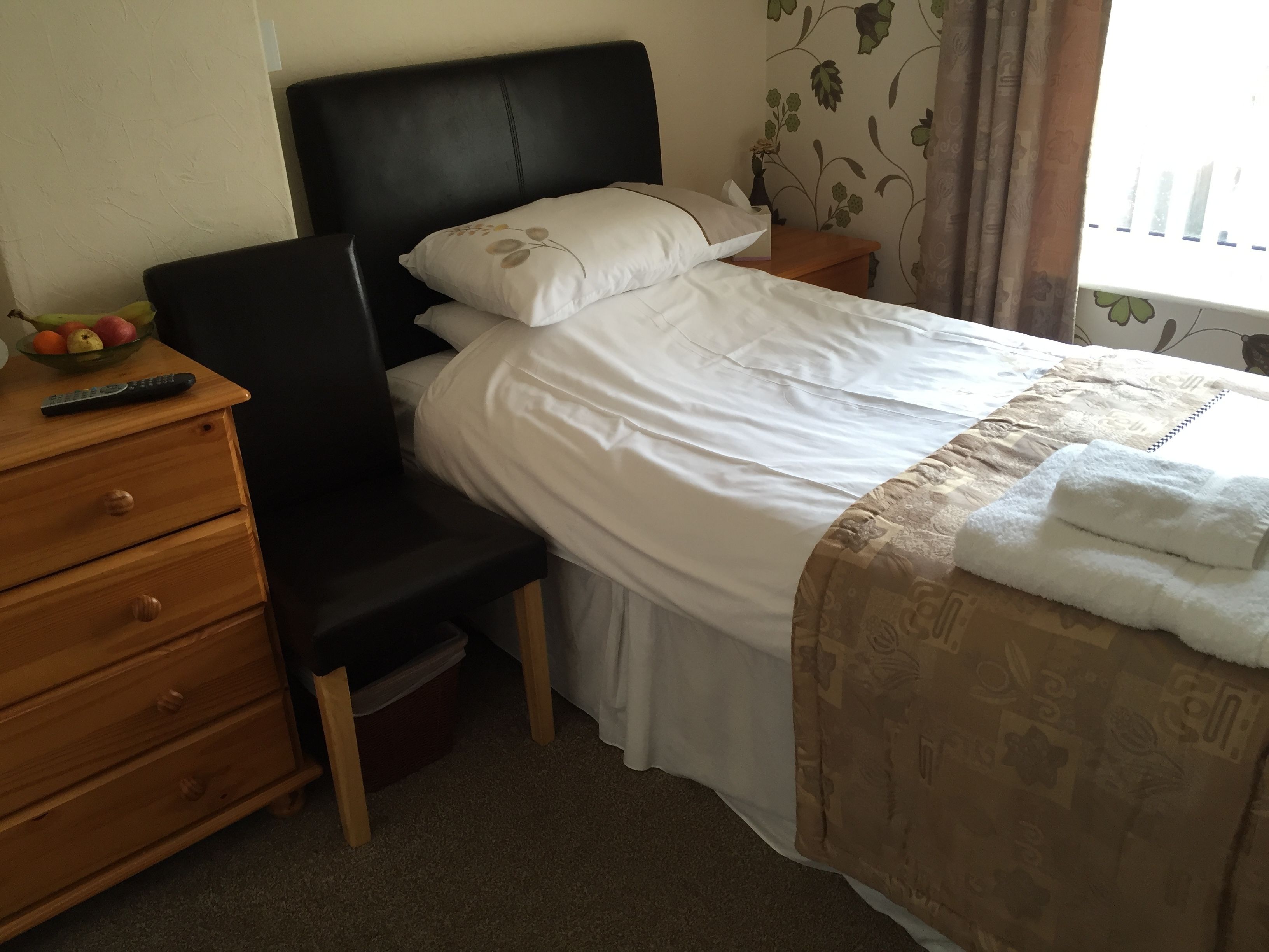 A neatly made bed with white linens, a wooden nightstand with a remote control, and a small wooden chair.