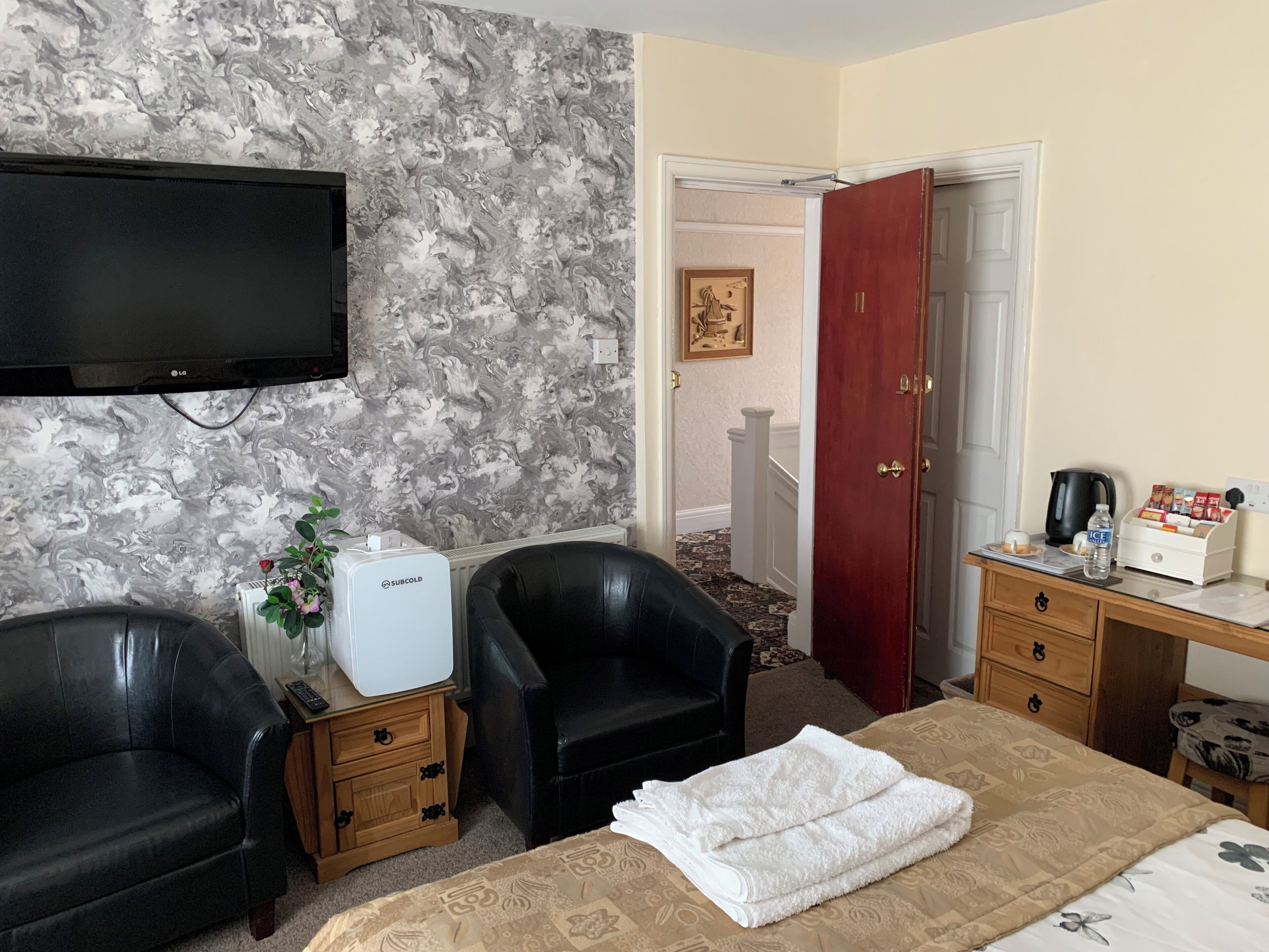 A hotel room with a marble-patterned wall, a flat-screen TV, two black leather chairs, a bed with a beige bedspread, a white towel, a Subcold refrigerator, a wooden nightstand, a wooden desk with a coffee maker, a water bottle, and a framed picture on the wall.