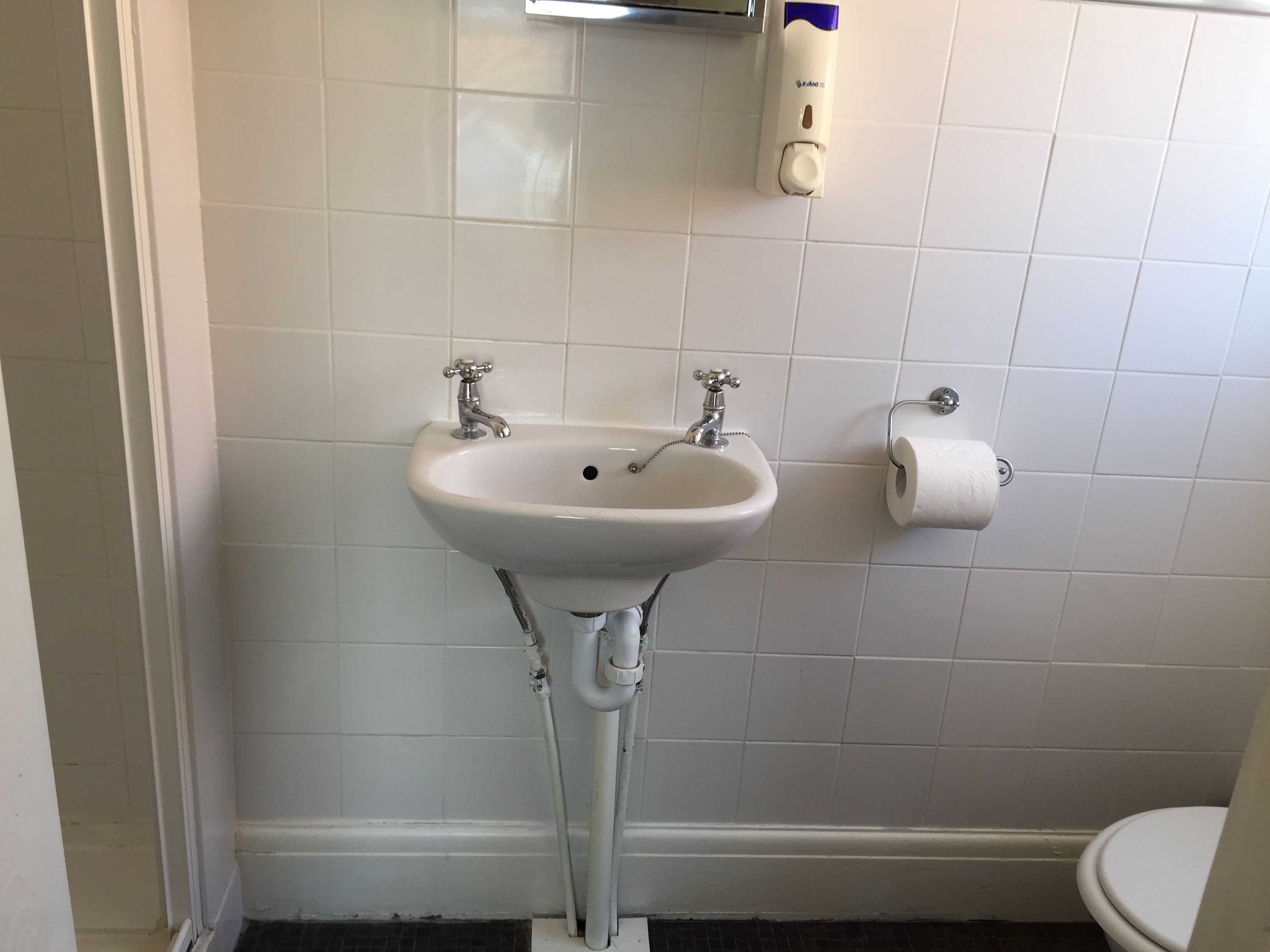 A bathroom with a white sink, two faucets, a toilet, and a paper towel dispenser.