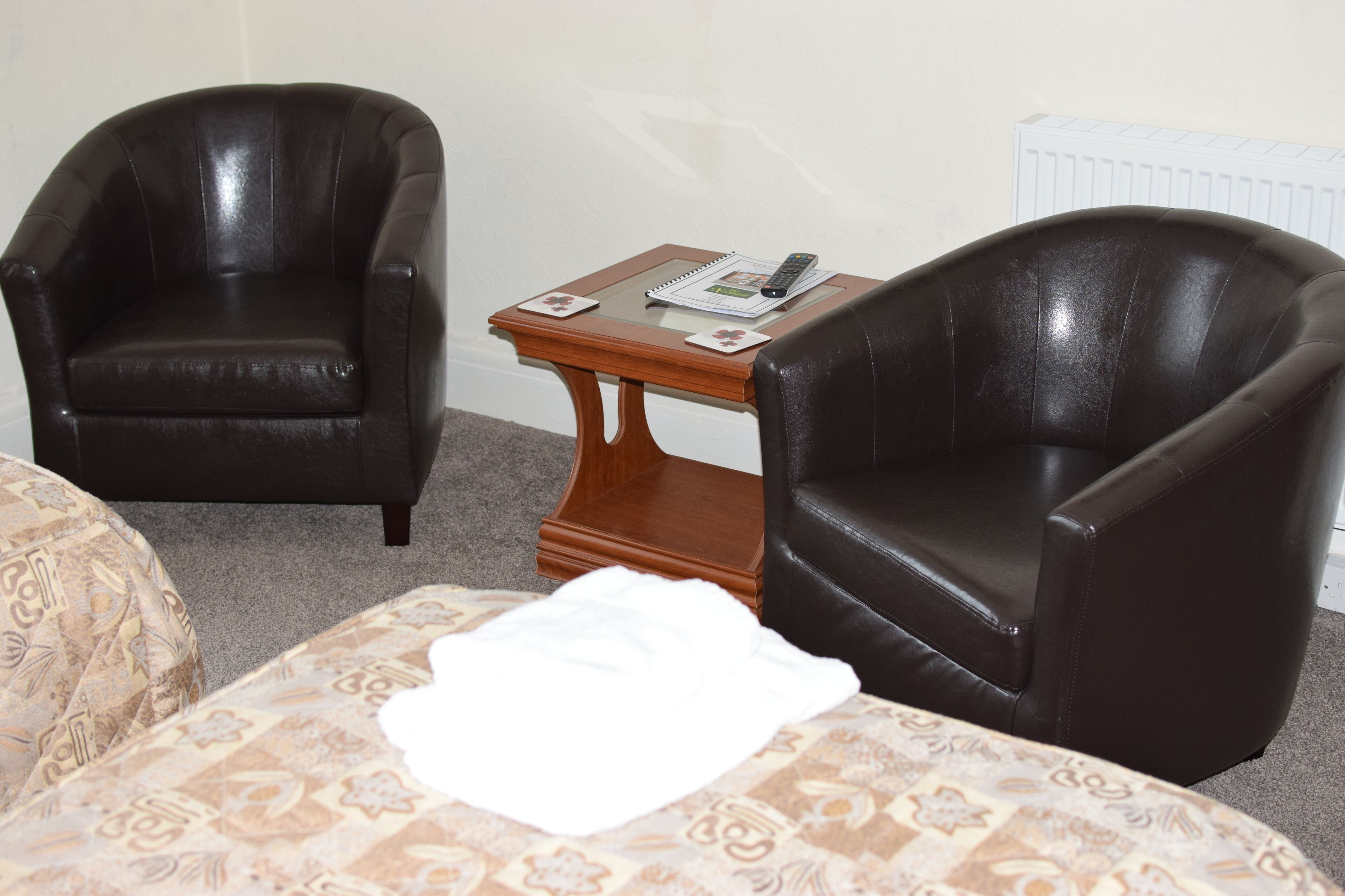 A room with two leather armchairs, a small wooden table with a remote control and a magazine, and a bed with a patterned bedspread.