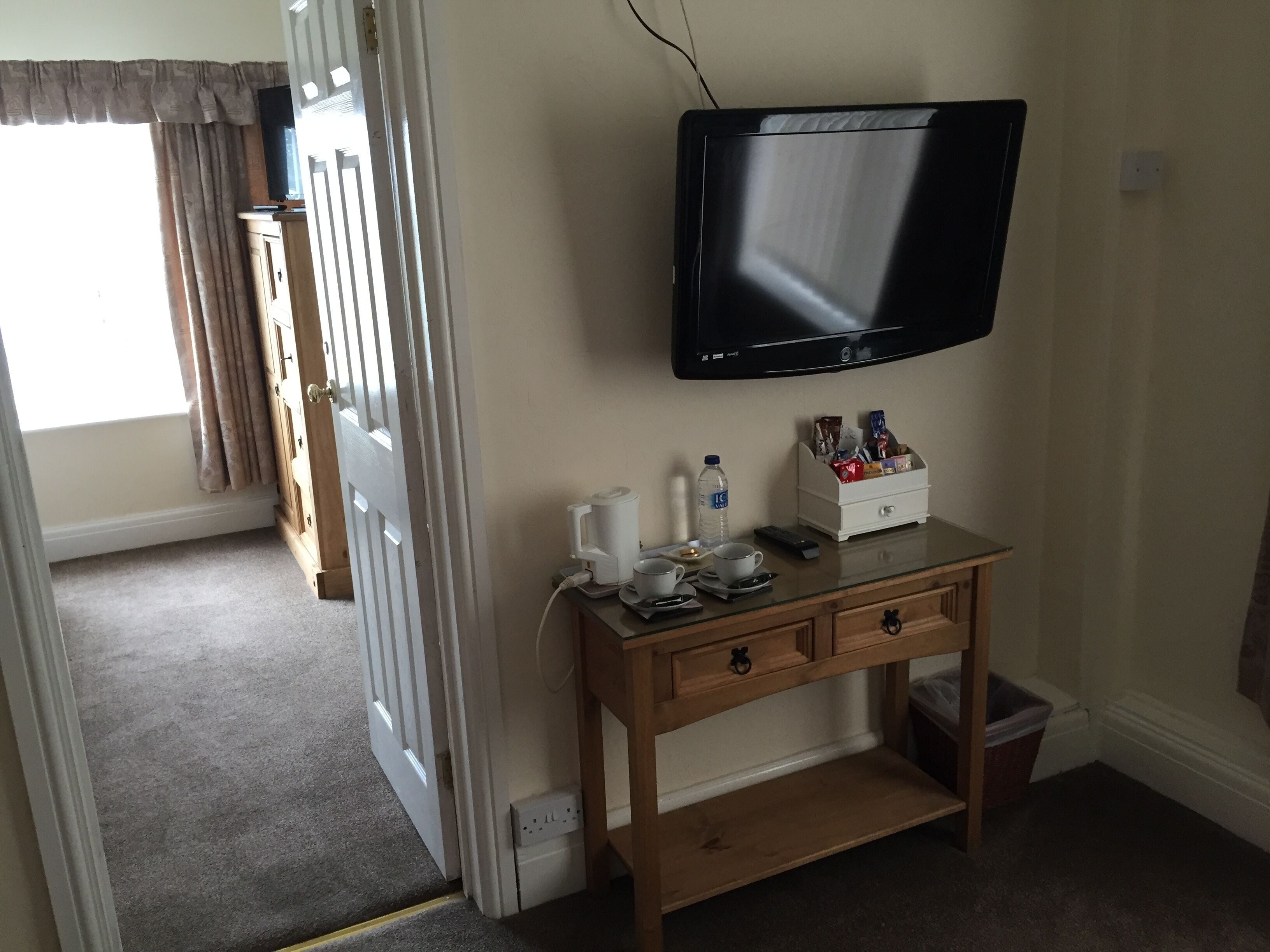 A hotel room with a television mounted on the wall, a wooden console table with a coffee maker, cups, and snacks, and a door leading to another room.