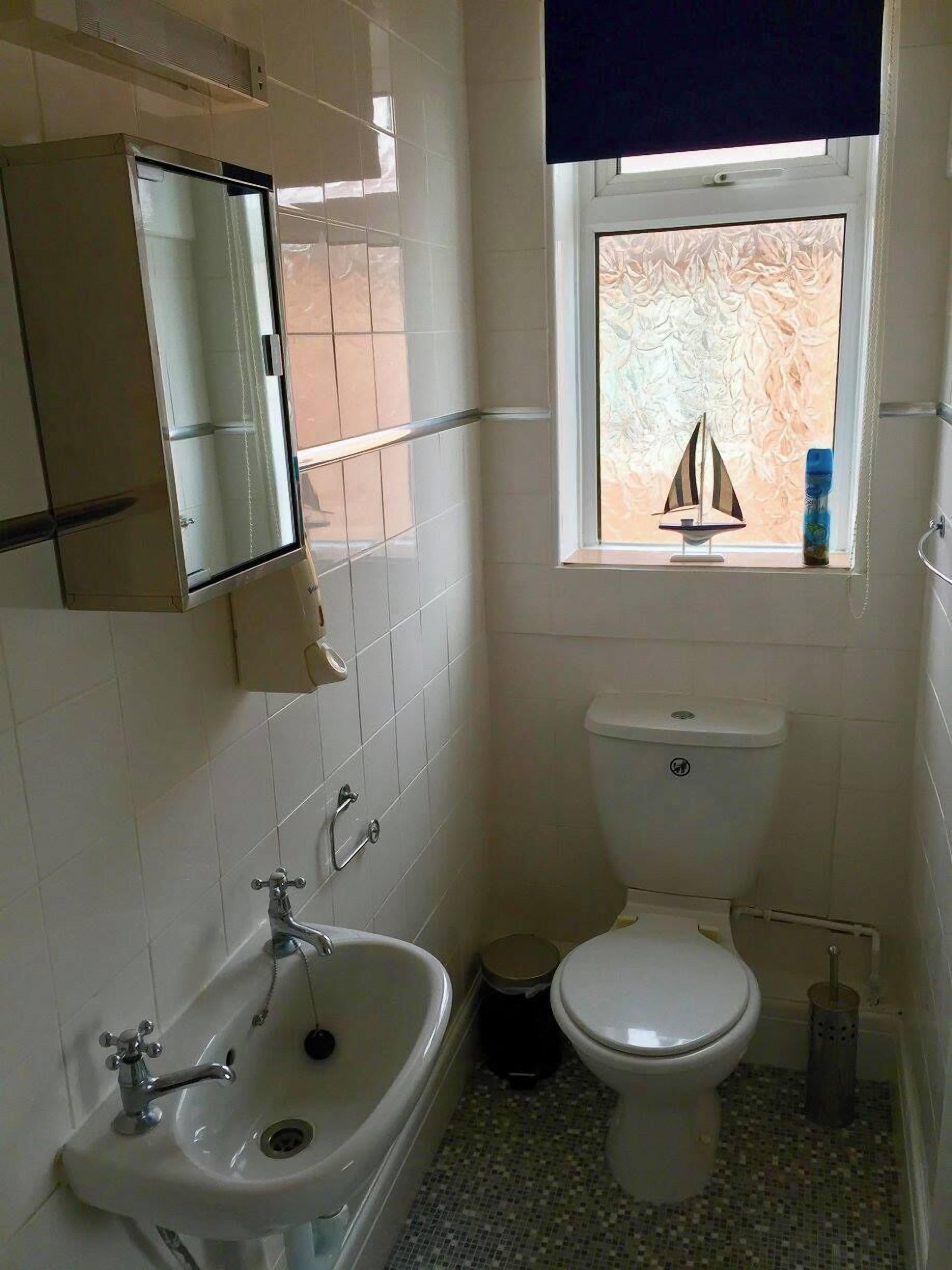 Casa de banho com lavatório branco, sanita e janela com um barco à vela decorativo.