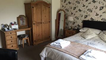 A bedroom with a bed, a wooden wardrobe, a desk with drawers, a chair, a mirror, and a nightstand with a lamp.