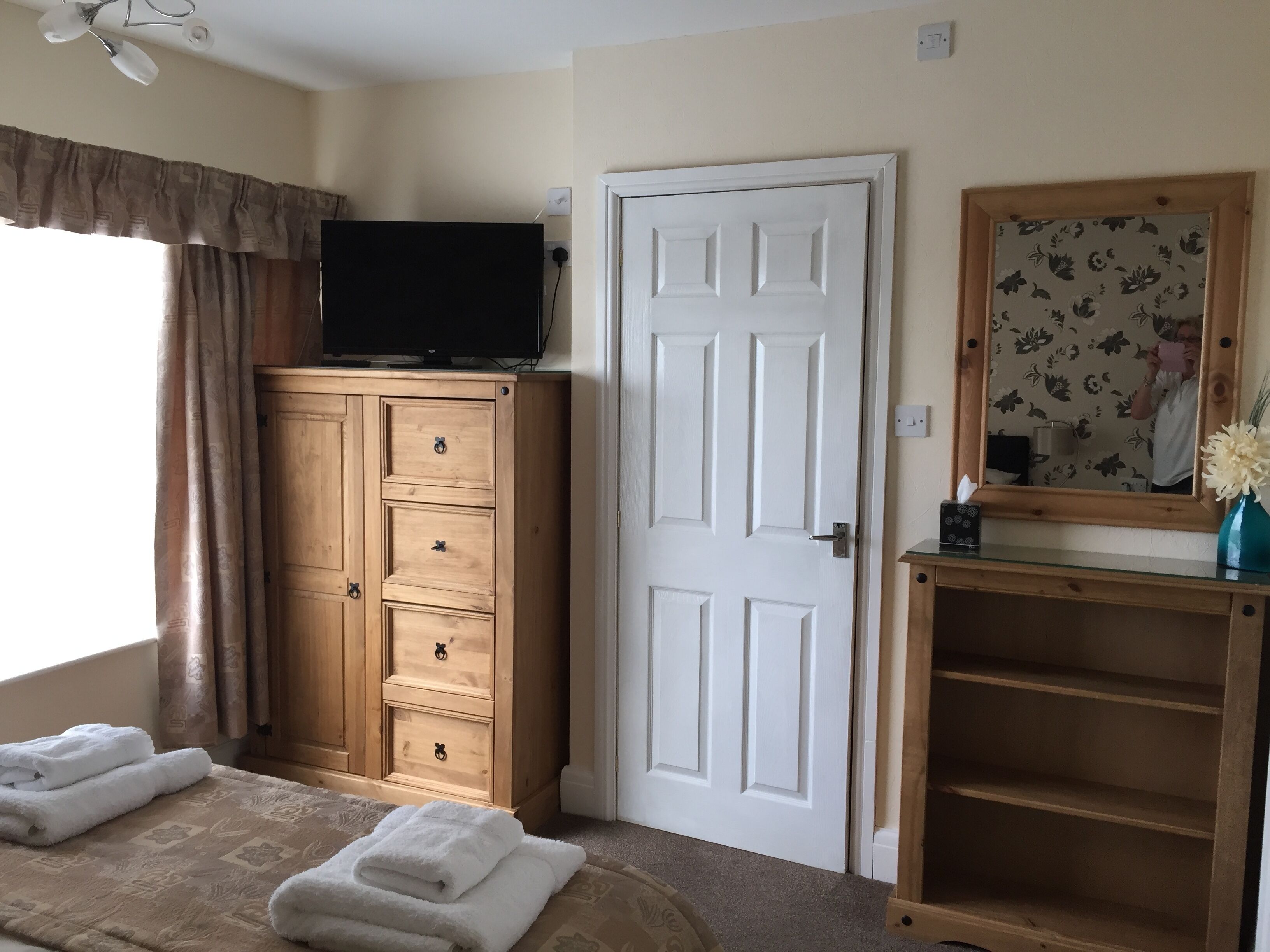 A bedroom with a wooden wardrobe, a white door, a television on top of the wardrobe, a bed with white towels, and a mirror with a floral frame.