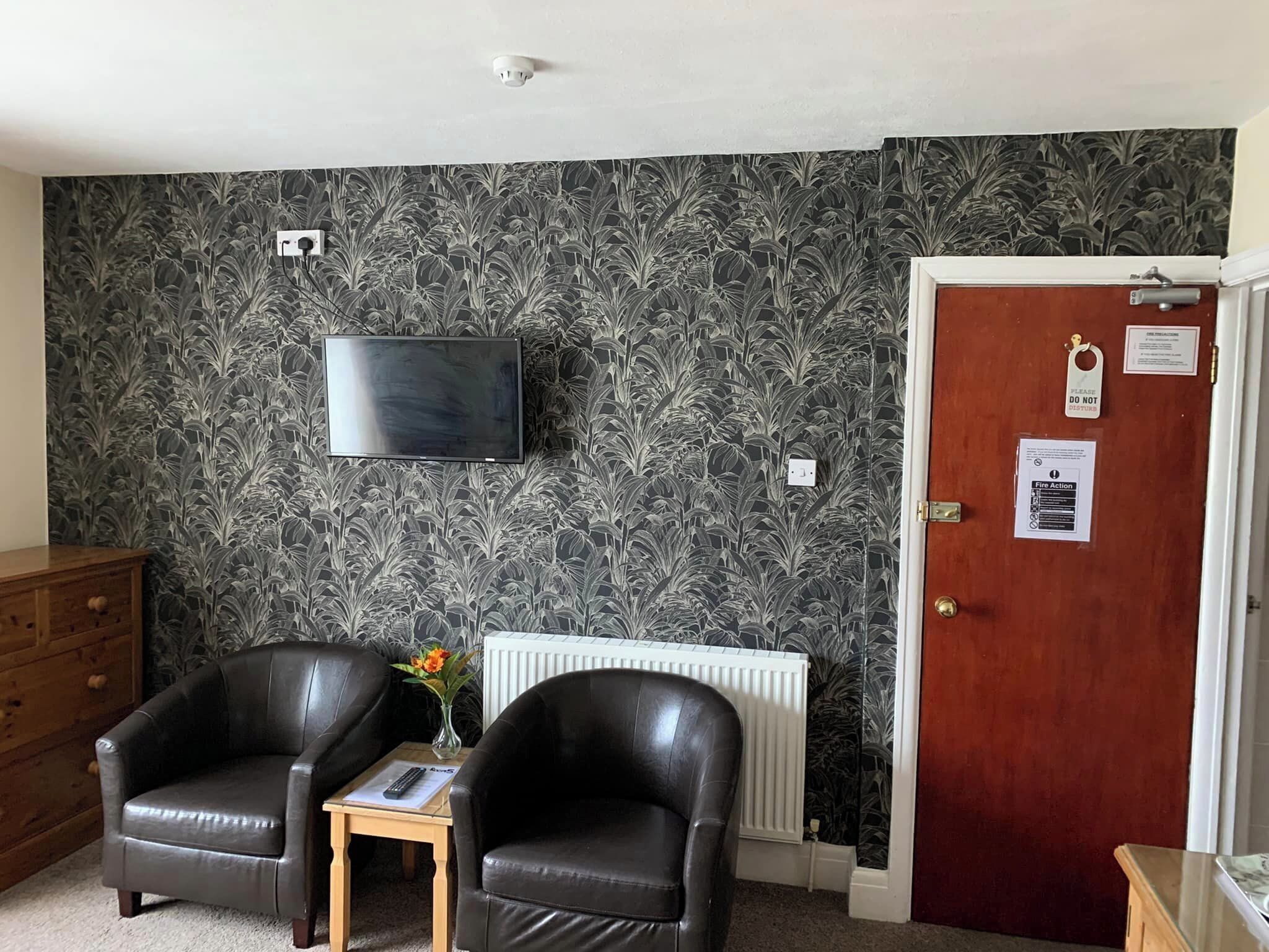 A room with a black leather sofa, a small wooden table with a vase, a radiator, a television mounted on the wall, and a door with a fire action sign.