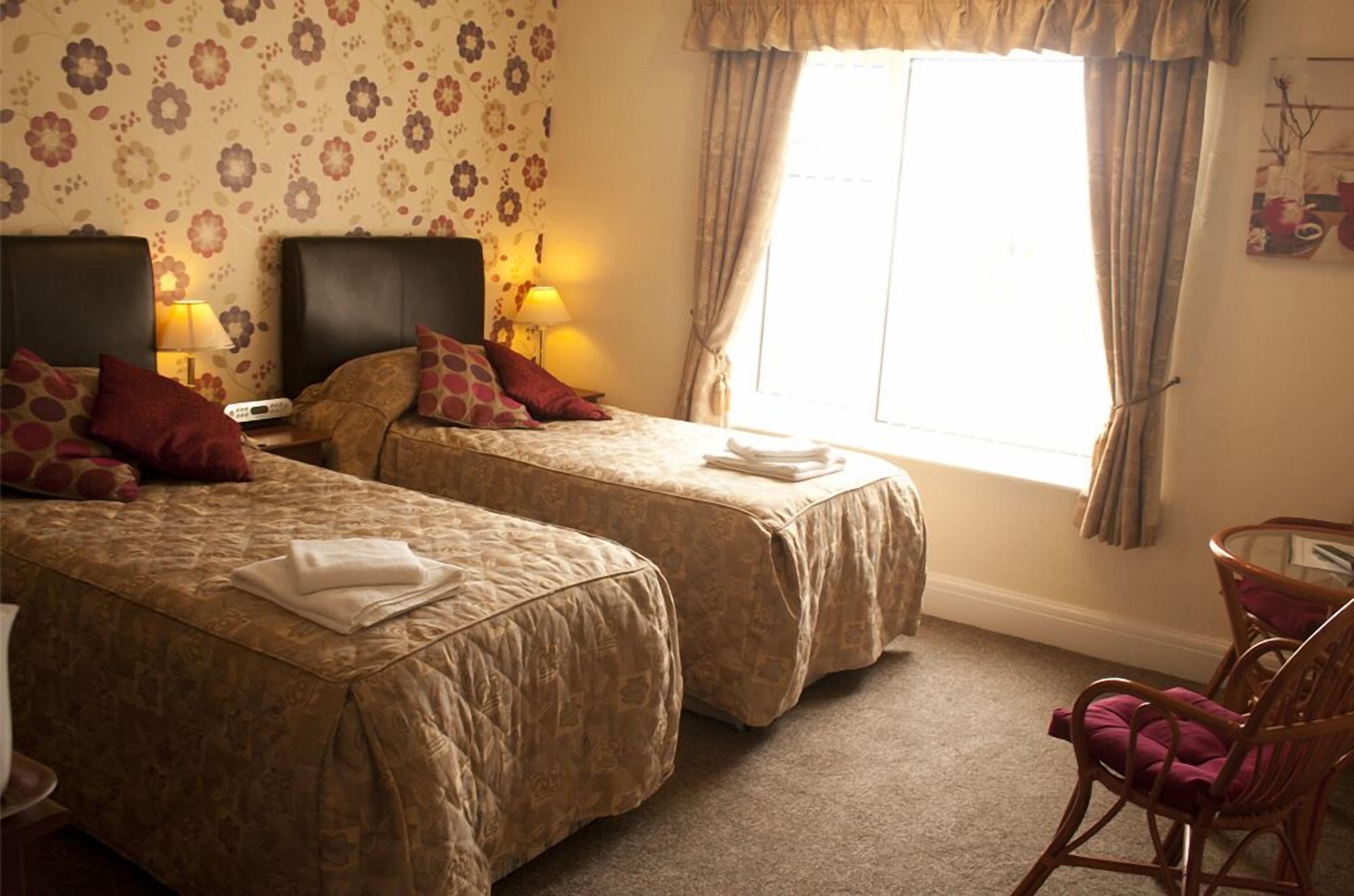A hotel room with two beds, each with a headboard, a small table, a chair, and a window with curtains.