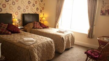 A hotel room with two beds, each with a headboard, a small table, a chair, and a window with curtains.