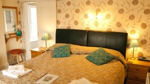 A neatly made bed with a patterned bedspread, two pillows, and folded towels. There are two bedside lamps, a nightstand, and a small stool. The room has floral wallpaper and a window with curtains. - The Northdale Hotel Ltd (Skegness)