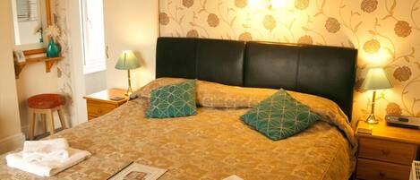 A neatly made bed with a patterned bedspread, two pillows, and folded towels. There are two bedside lamps, a nightstand, and a small stool. The room has floral wallpaper and a window with curtains.