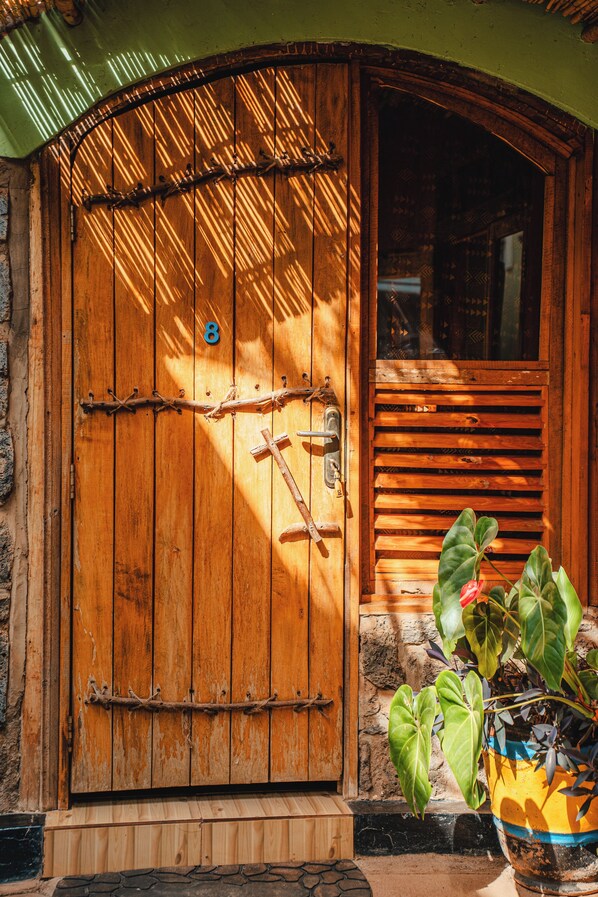 A wooden door with a number '8' and a rope tied across it, a window with horizontal slats, and a potted plant with broad leaves. - Yambi Guesthouse (Kigali)
