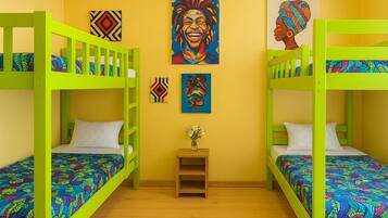 A room with two bunk beds, each with a colorful patterned bedspread. There are two paintings on the wall, one depicting a joyful figure and the other a profile of a person. A small wooden nightstand with a vase of flowers is in the center.
