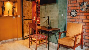 A room with a brick wall, a television mounted on it, a table with a chair, and a shelf with decorative items.