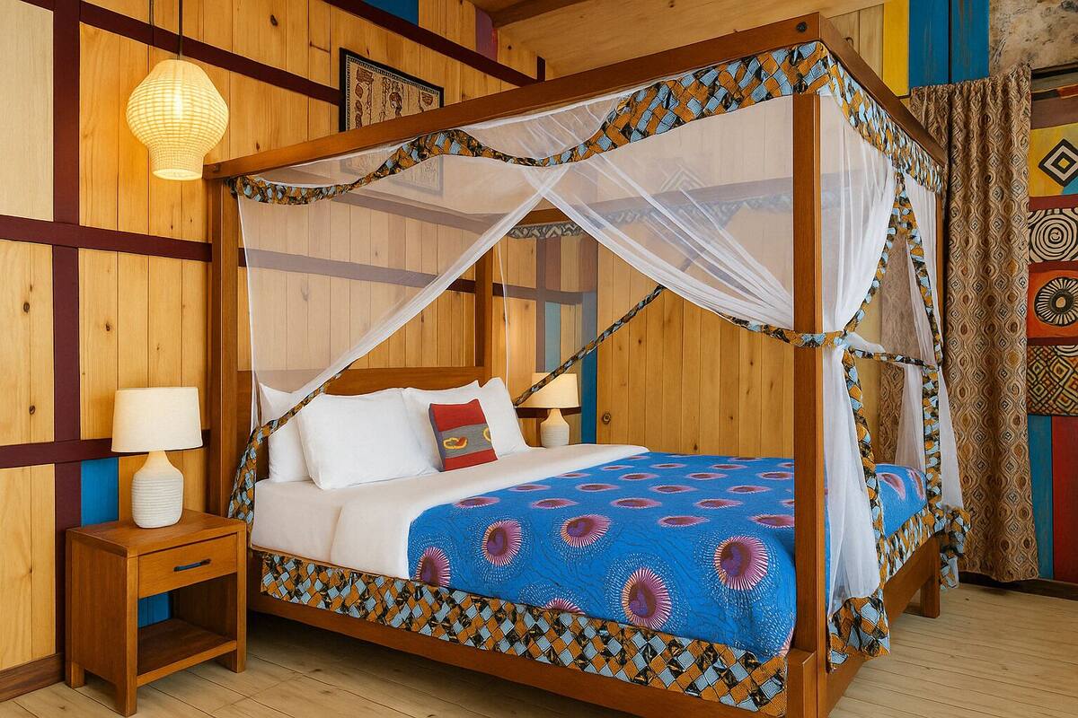 A wooden four-poster bed with a blue patterned bedspread and white pillows, a bedside table with a lamp, and a hanging light fixture.
