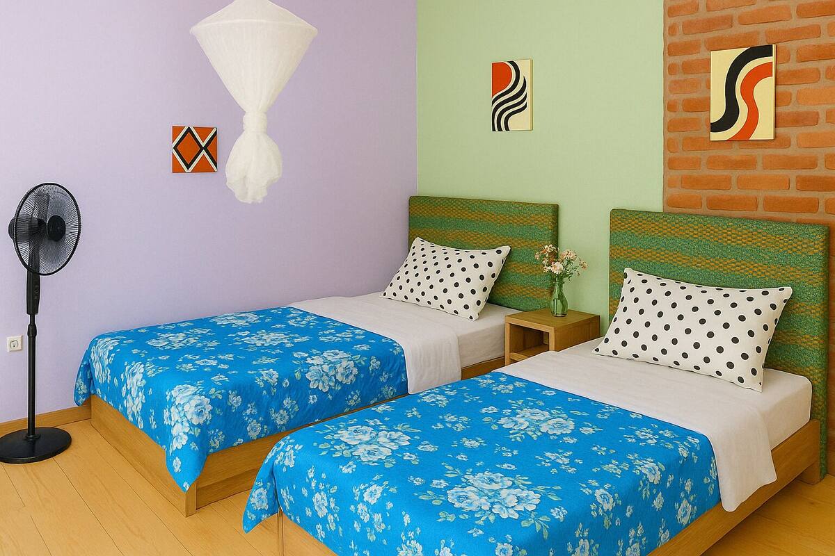 A room with two beds, each with a blue floral bedspread, a standing fan, and a brick wall with artwork.