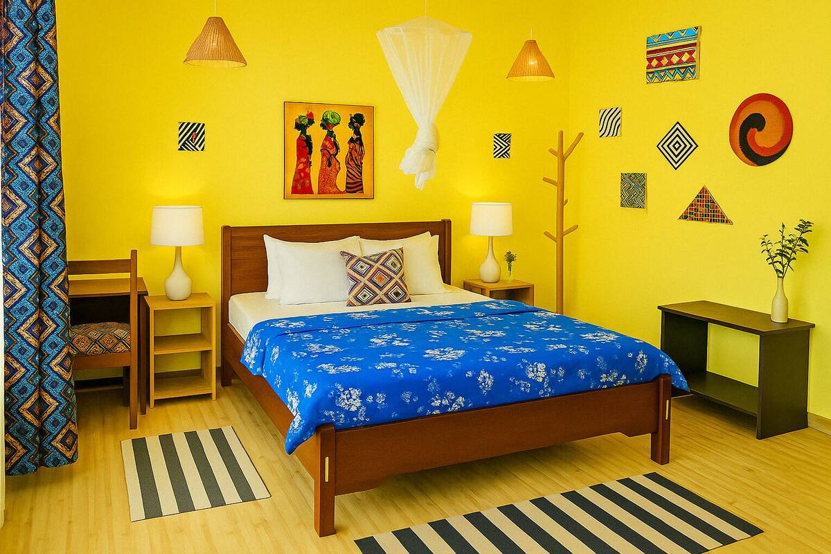 A room with a bed, two bedside tables, a nightstand, a chair, a hanging lamp, a floor lamp, a wall with decorative patterns, and a small table with a vase.