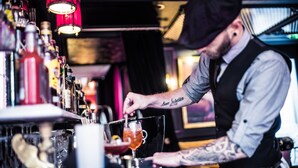 A bartender preparing a drink behind a bar with various bottles and glasses. - Hôtel de JoBo (Paris)