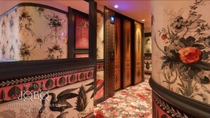 A hotel corridor with floral wallpaper, a wooden bench, and a door with a red frame. - Hôtel de JoBo (Paris)