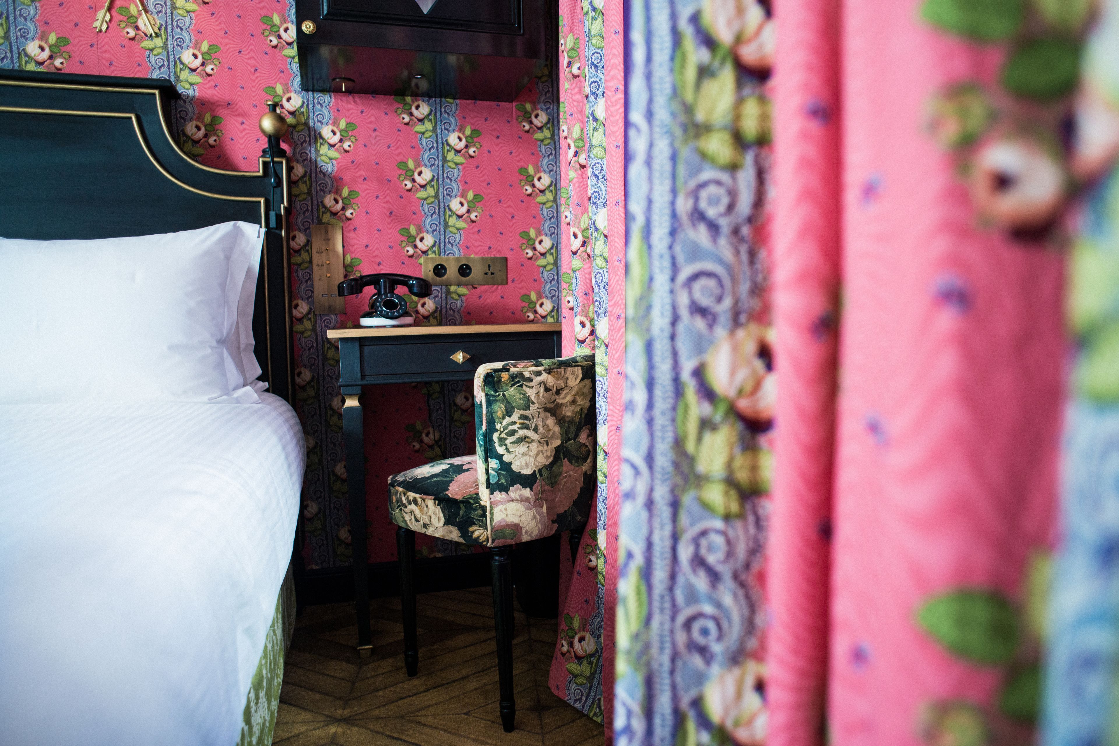 Une chambre avec un lit, un bureau et une chaise. Les murs sont recouverts d’un papier peint à motifs floraux et il y a un rideau rose à la fenêtre.