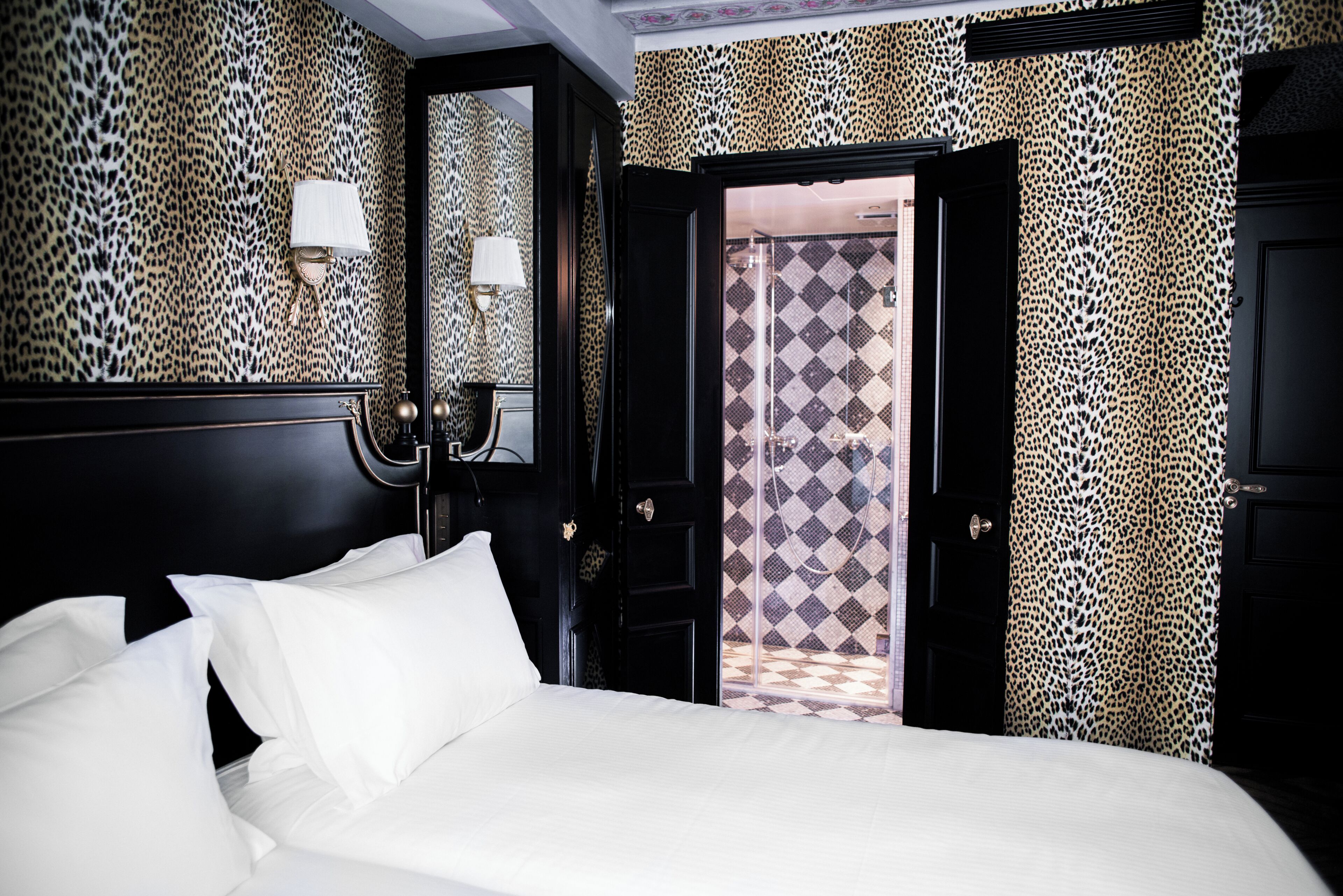 A hotel room with a bed, a nightstand, a lamp, a mirror, and a door leading to a bathroom with patterned tiles.