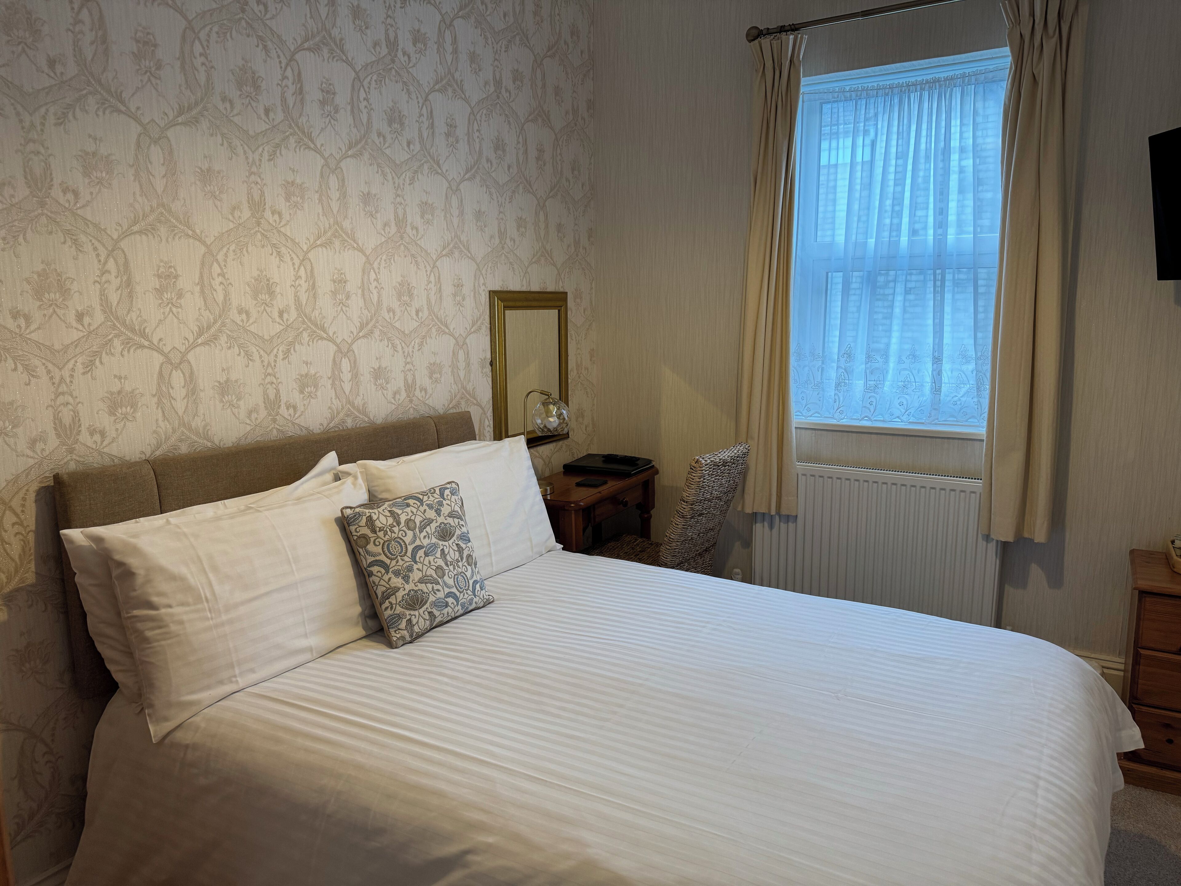 A neatly made bed with white bedding and patterned pillows, a bedside table with a lamp, a chair, and a window with curtains.