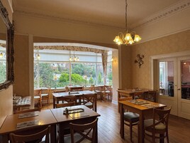 Zona pranzo con tavoli e sedie in legno, ampie finestre con tende e un lampadario.