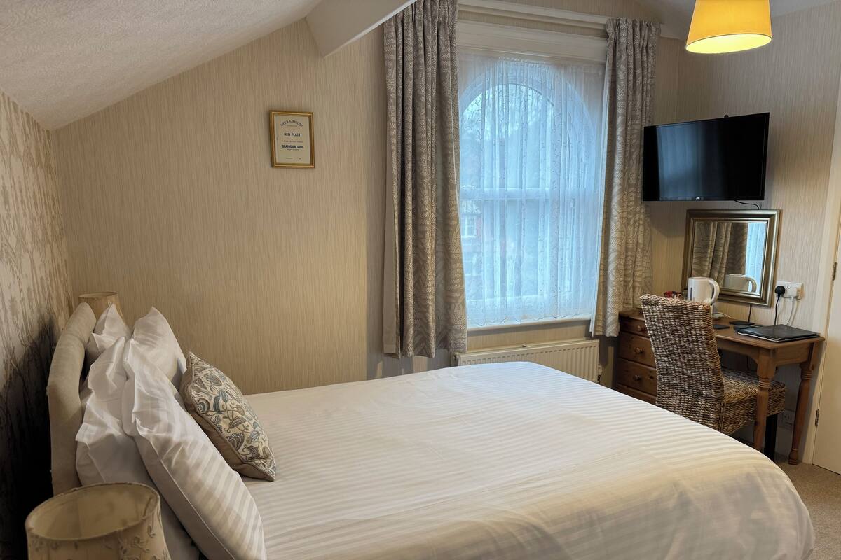 A neatly made bed with a white comforter and pillows, a wooden nightstand with a lamp, a wicker chair, a television, and a framed picture on the wall.