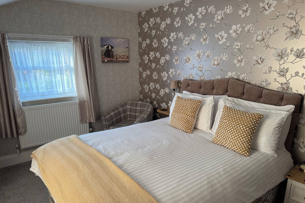 A neatly made bed with patterned pillows, a plaid chair, and a floral wallpaper.