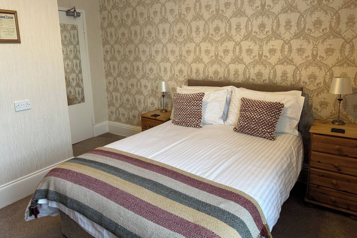 A neatly made bed with patterned pillows, a bedside table with a lamp, and a wall with a floral wallpaper pattern.