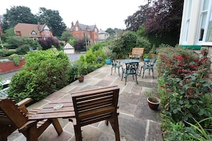A patio with wooden furniture, potted plants, and a view of a red brick building in the background.
